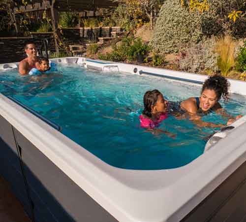 A group of people enjoy a swim in a backyard pool, surrounded by plants and outdoor furniture under a clear sky.