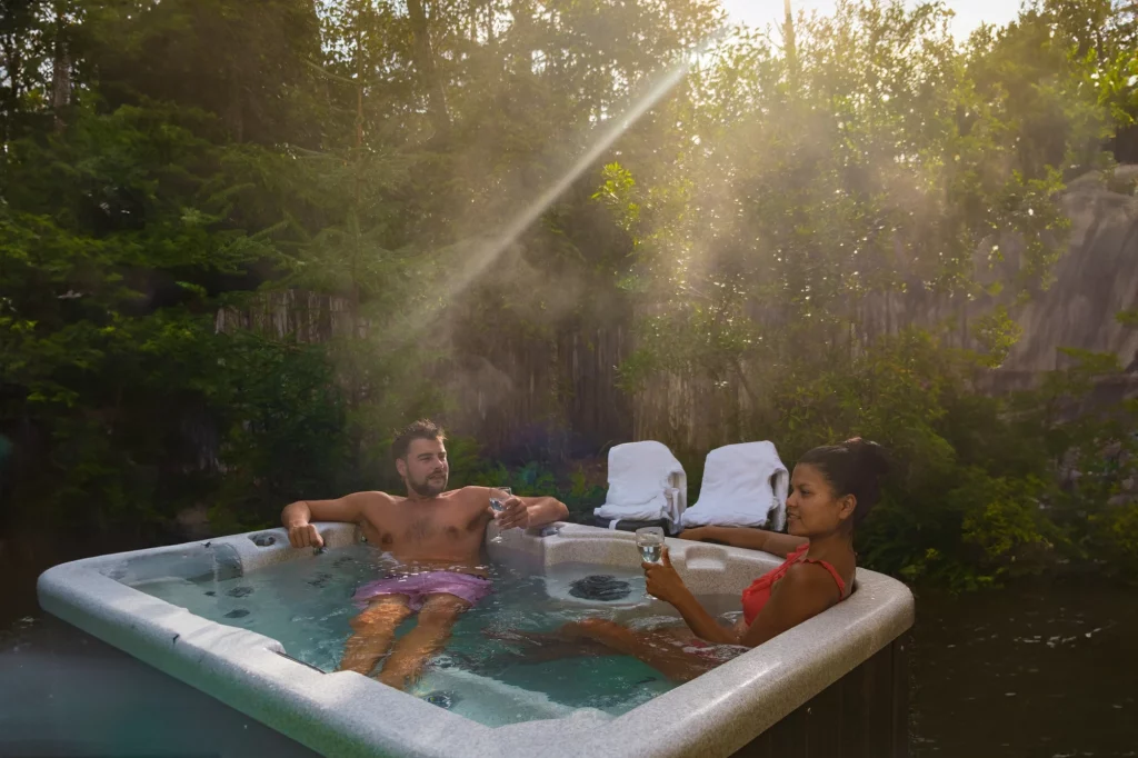 Two people relax in an outdoor hot tub, surrounded by lush greenery, with sunlight streaming softly through the trees.