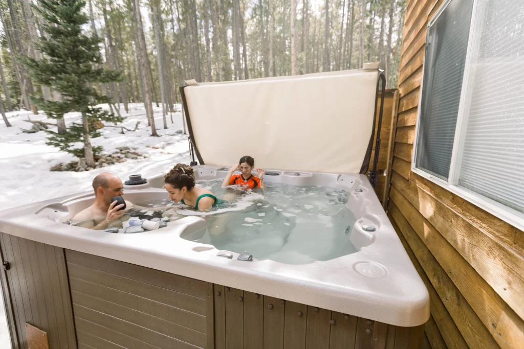 Three people relaxing in a hot tub outside a wooden cabin, surrounded by snowy forest. They are enjoying a cold weather retreat.