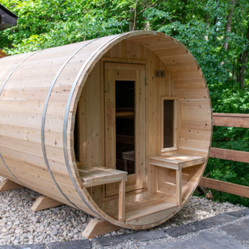 A wooden barrel sauna sits outdoors on a gravel base, surrounded by lush green foliage, with seating areas near the entrance.