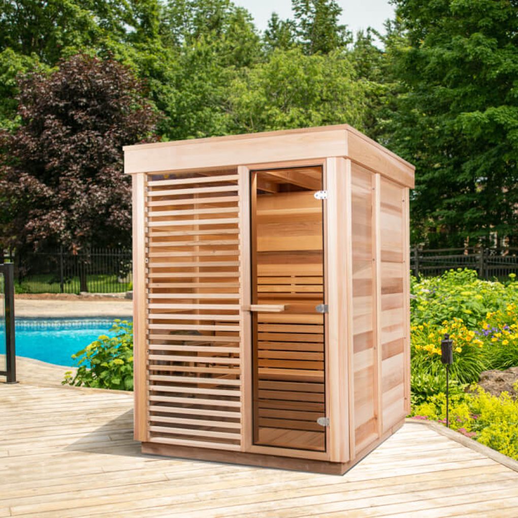 A small wooden sauna sits on a deck beside a pool, surrounded by lush greenery and vibrant flowers under a clear sky.