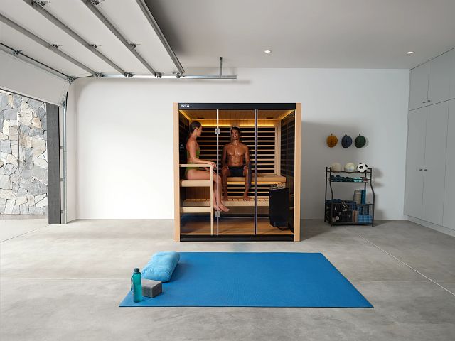 Two people sit in a modern home sauna within a spacious garage, featuring exercise equipment and a blue yoga mat on the floor.