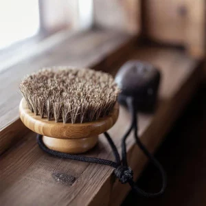 A wooden body brush with natural bristles sits on a rustic windowsill, accompanied by a black cord for hanging storage.