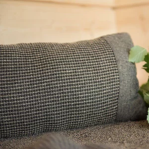 Textured gray cushion on checkered fabric surface, with a glimpse of green leaves nearby, placed against a light-colored wooden wall background.