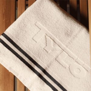 White towel with "TYLO" embossed, featuring two dark stripes on wooden slats. Cozy, minimalist design suitable for a spa or sauna setting.