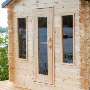 A small wooden cabin with three glass windows overlooks a serene lake, surrounded by lush greenery and distant trees under a bright sky.
